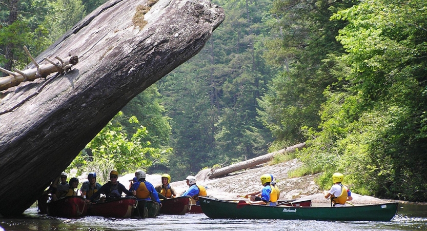 People sit in canoes resting in the shade of a large rock.